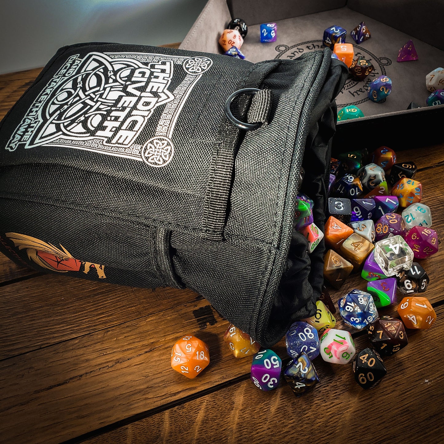 Black dice bag with white design on a wooden surface surrounded by colorful dice. Design reads "The dice giveth and the dice taketh away".