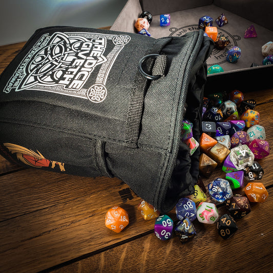 Black dice bag with white design on a wooden surface surrounded by colorful dice. Design reads "The dice giveth and the dice taketh away".